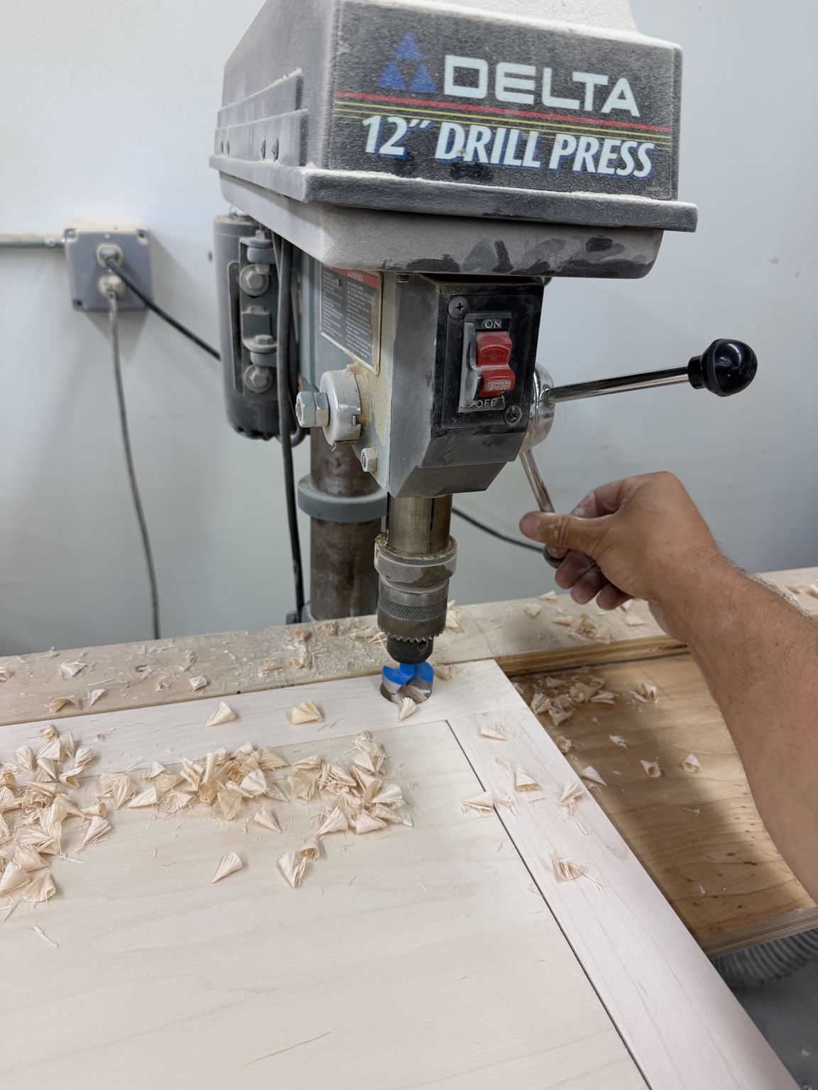 Wood panel at a drill press with pencil layout marks and fresh wood shavings