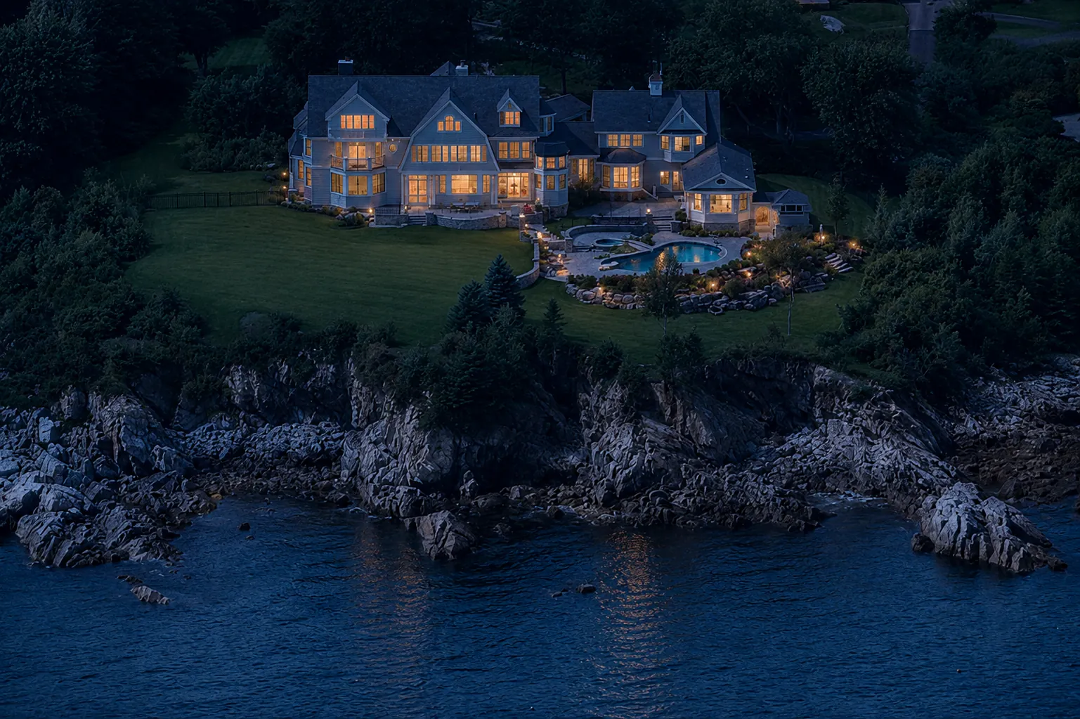 Shingle-style coastal estate on Shore Road in Cape Elizabeth glowing above the rocky Atlantic shoreline at dusk
